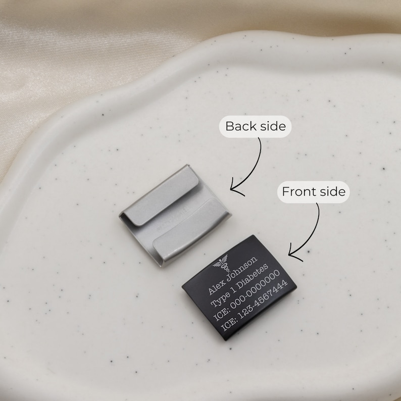 K&ouml;nnte beinhalten: Ein schwarzes medizinisches Anh&auml;nger aus Metall mit dem Text "Alex Johnson Typ 1 Diabetes ICE: 000-0000000 ICE: 123-4567444" und einem medizinischen Symbol. Der Anh&auml;nger ist an einem silbernen Metallclip befestigt.