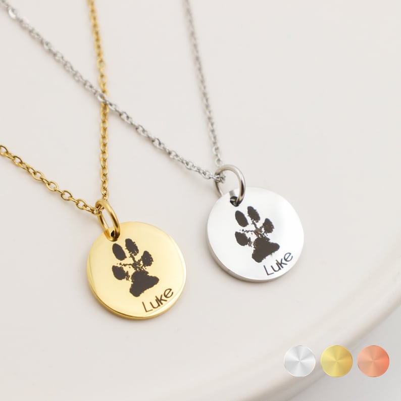 May include: Two gold and silver necklaces with round pendants featuring a paw print and the name "Luke" engraved on each. The necklaces are on a white background with three small circles showing the different metal options: silver, gold, and rose gold.