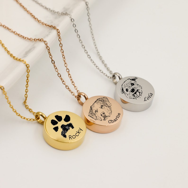 3 round cremation urn necklaces for pet ashes resting on a white surface. The gold necklace is engraved in black with the paw print and the name curved around the edge under the paw. The silver and rose gold have the face of the dog and the name