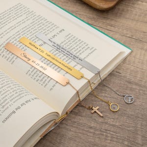 May include: Three gold, silver and rose gold bookmarks with chains and charms. The bookmarks are engraved with inspirational quotes and a name and date. The charms include a cross, a compass and a small tree.