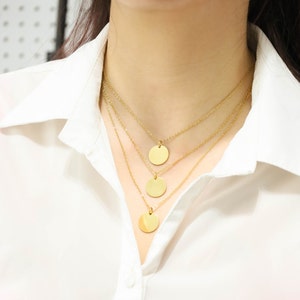 May include: A layered gold necklace with three round pendant charms. The necklace is worn by a person with a white shirt.