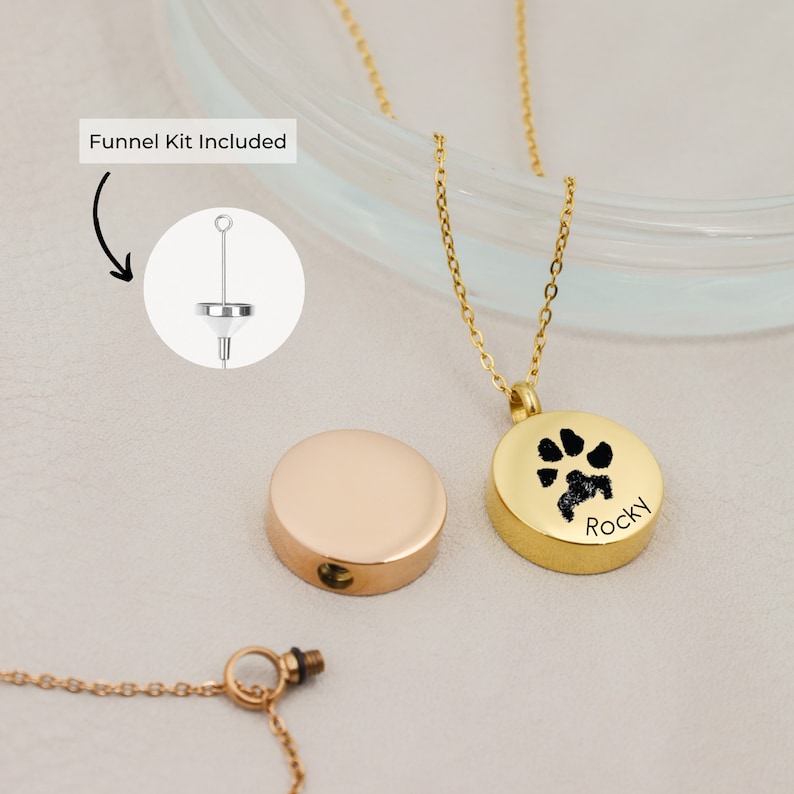 May include: Gold tone stainless steel cremation necklace with a paw print charm. The charm has the name "Rocky" engraved on it. The necklace comes with a funnel kit for filling the pendant.