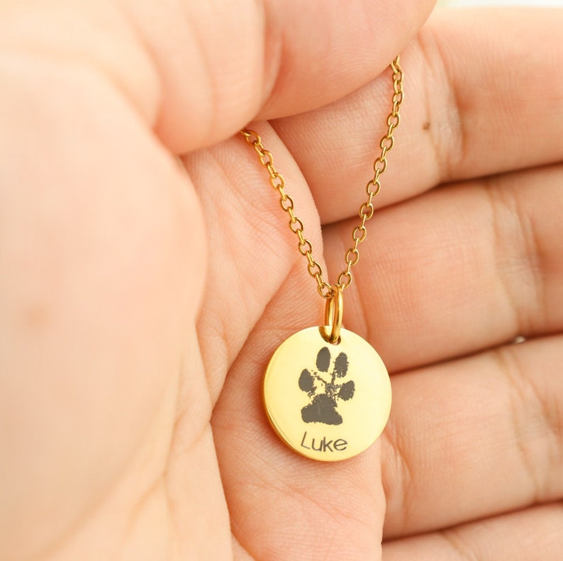 May include: Gold necklace with a round pendant featuring a black paw print and the name "Luke" engraved on it.