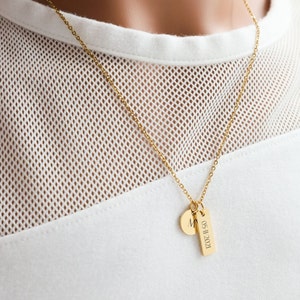 May include: A gold chain necklace with a small round pendant engraved with the letter "M" and a longer rectangular pendant engraved with the date "05.11.2021".