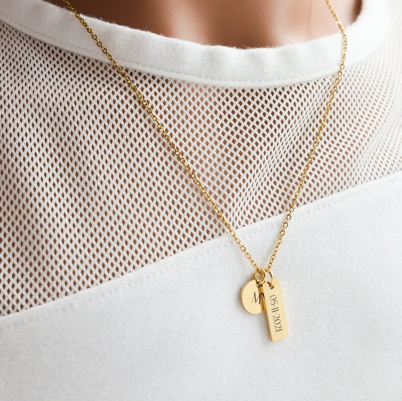 May include: A gold chain necklace with a small round pendant engraved with the letter "M" and a longer rectangular pendant engraved with the date "05.11.2021".
