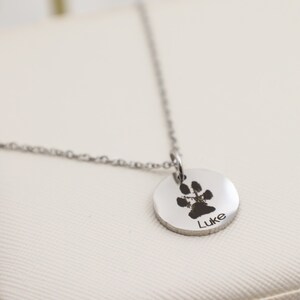 May include: Silver necklace with a round pendant featuring a paw print and the name "Luke" engraved on it.