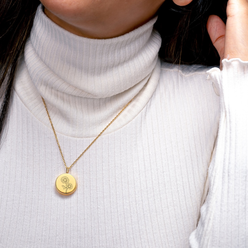 May include: A gold chain necklace with a round pendant featuring a delicate engraved sunflower design. The necklace is worn by a person wearing a white turtleneck sweater.