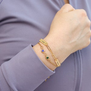 May include: Gold chain bracelet with two personalized nameplate charms, one with the name "Evans" and the other with the name "Patrick". The bracelet also features blue and green gemstones.