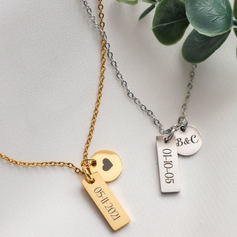 May include: Two gold necklaces with personalized pendants. One necklace has a small heart charm and a rectangular pendant with the date "05-11-2021" engraved on it. The other necklace has a round pendant with the initials "B&C" engraved on it and a rectangular pendant with the date "01-10-05" engraved on it.