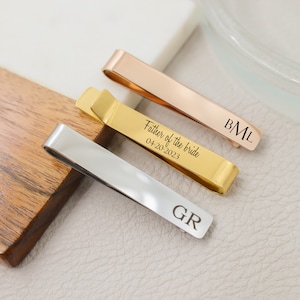 May include: Three tie bars with different engravings. The top tie bar is rose gold with the initials "BML" engraved on it. The middle tie bar is gold with the text "Father of the bride 04-20-2023" engraved on it. The bottom tie bar is silver with the initials "GR" engraved on it.