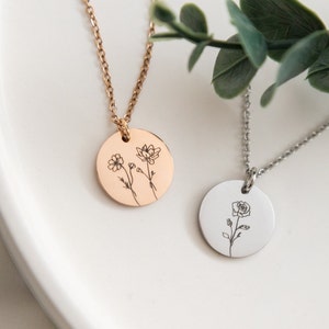 May include: Two delicate necklaces with round pendants featuring engraved floral designs. One pendant has a rose and the other has two smaller flowers. The necklaces are on a white surface.