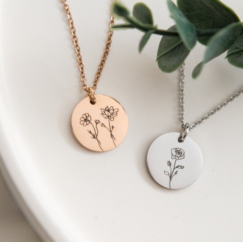 May include: Two delicate necklaces with round pendants featuring engraved floral designs. One pendant has a rose and the other has two smaller flowers. The necklaces are on a white surface.