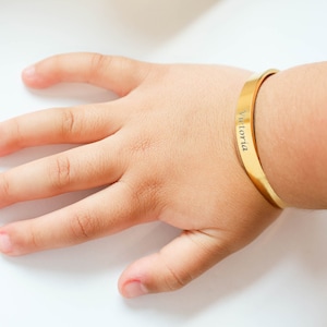 May include: A gold-coloured bracelet with the name "Victoria" engraved on it. The bracelet is worn on a child's wrist.