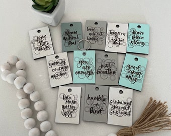 May include: A set of 12 wooden tags with inspirational quotes and a beaded garland with a tassel. The tags are painted in shades of gray, white, and blue with black lettering. The quotes include "Dream without fear", "Love without limits", "Tomorrow is another day", "Brave fierce strong", "Serenity courage wisdom", "You are enough", "Just breathe", "Good vibes only", "Live more worry less", "Humble or kind", and "She believed she could so she did".