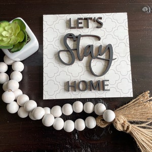 May include: White wooden beads with a brown tassel, a white planter with a green succulent, and a white sign with black lettering that reads "Let's Stay Home".