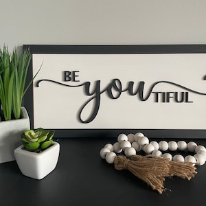 Könnte beinhalten: Ein schwarzes gerahmtes Schild mit weißem Hintergrund und dem Text "BE YOU TIFUL" in schwarzer Kursivschrift. Das Schild steht auf einer schwarzen Oberfläche mit einem weißen Topf mit grünen Pflanzen und einem weißen Topf mit grünen Sukkulenten.
