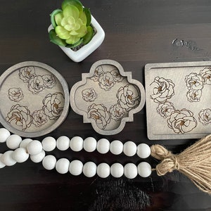 May include: A selection of decorative wooden coasters in round, quatrefoil, and square shapes. Each coaster has a laser-etched floral design. A white bead garland and a small potted succulent are also present.