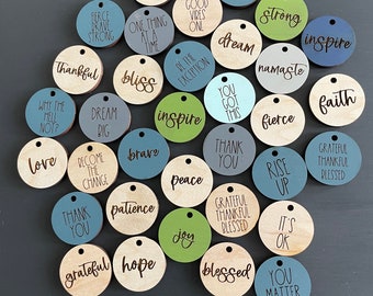 Puede incluir: Una colección de círculos de madera con palabras y frases inspiradoras, como "gracias", "amor", "paz", "agradecido", "bendecido", "esperanza", "alegría", "inspirar", "sueño", "fe", "fuerte", "feroz", "valiente", "levantarse", "está bien", "tú importas", "querer", "namaste", "ser la excepción", "¿por qué no?", "una cosa a la vez", "convertirse en el cambio", "buenas vibraciones solamente", "tú tienes esto", "soñar en grande", y "paciencia".