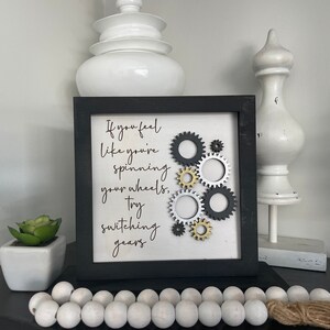 Pode incluir: Arte de parede emoldurada em preto com fundo branco e o texto "If you feel like you're spinning your wheels, try switching gears." O texto está em fonte de letra cursiva preta. Há várias engrenagens prateadas e douradas no fundo branco.