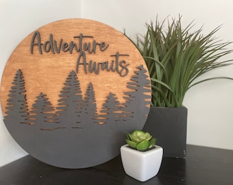 May include: A round wooden sign with the text "Adventure Awaits" in black lettering. The sign is painted black on the bottom half and features a silhouette of a forest with pine trees.