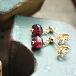 May include: A pair of gold earrings with small, faceted, dark red gemstones. The earrings have a simple, minimalist design.