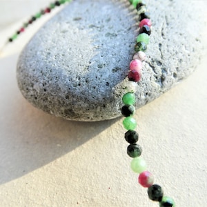 May include: A strand of gemstone beads in black, green, and pink. The beads are faceted and have a shiny finish. The beads are strung on a clear thread and are lying on a grey stone.