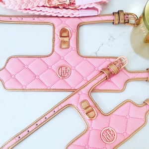 Two pink quilted dog harnesses with gold trim and a gold monogram. The harnesses have a gold buckle and a gold D-ring. A pink braided rope leash is also included.