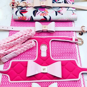 A set of pink and white dog collars and harnesses with a quilted design and pink bows. The collars and harnesses feature a gold metal logo with the letters "ld".