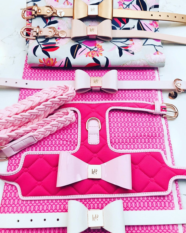 A set of pink and white dog collars and harnesses with a quilted design and pink bows. The collars and harnesses feature a gold metal logo with the letters "ld".