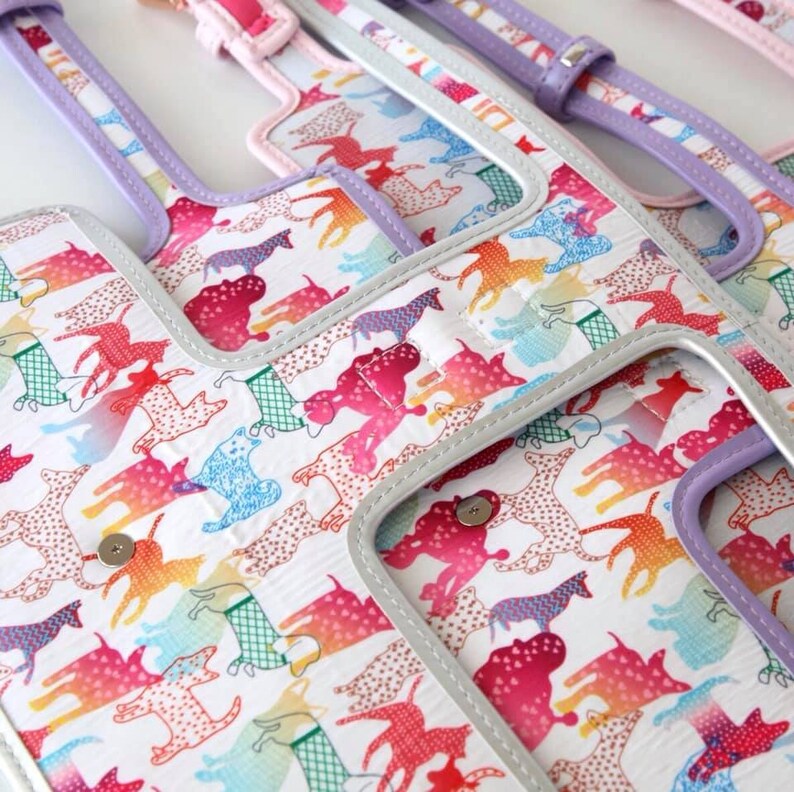 A close-up of a white fabric with a repeating pattern of colourful dogs. The fabric is trimmed with a silver and lavender coloured trim.