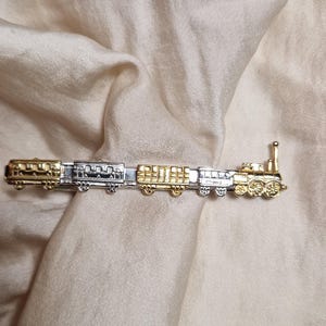Könnte beinhalten: Eine gold- und silberfarbene Krawattenklammer in Form eines Zuges. Die Klammer zeigt eine Lokomotive und mehrere Waggons, alle mit aufwendigen Details. Das Accessoire ist dazu bestimmt, eine Krawatte an Ort und Stelle zu halten und verleiht einen Hauch von Vintage-Charme.