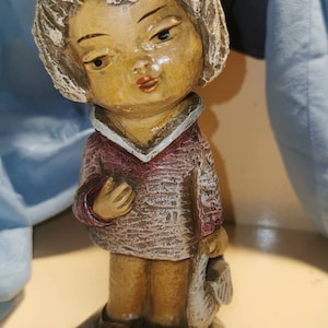 Puede incluir: Una figurita de un niño con cabello blanco, que lleva una camisa a rayas marrones y rosas y sostiene un pájaro blanco. La figurita está de pie sobre una base hexagonal.