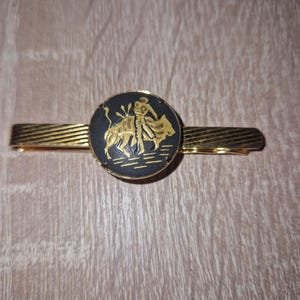 May include: Gold-toned tie clip with a circular black and gold emblem featuring a matador and bull. The clip has a textured design. A classic accessory for formal wear.