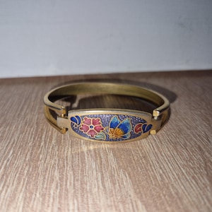 Puede incluir: Un brazalete de oro con un diseño de esmalte colorido que presenta una mariposa y flores. El diseño está sobre un fondo morado con acentos rojos, azules, amarillos y verdes.