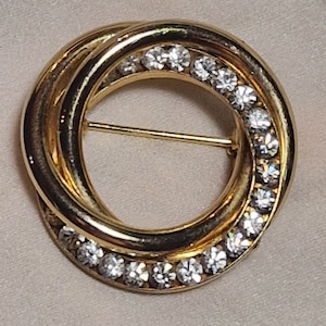 May include: A gold-toned brooch with a circular design. The brooch is made of three intertwined circles, each with a row of clear rhinestones.