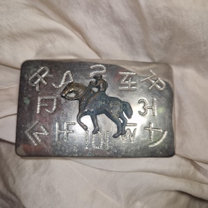 Puede incluir: Hebilla de cinturón rectangular plateada con un relieve en relieve de una persona montando a caballo. La hebilla tiene símbolos y letras grabados en los bordes. El jinete y el caballo son de un color más oscuro y oxidado.