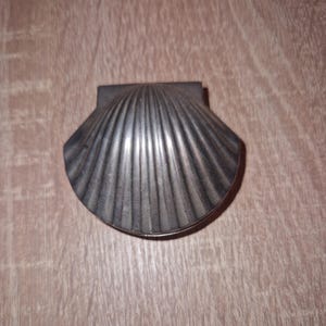 以下が含まれることがあります： 銀色の貝殻型の小物入れ。箱にはヒンジ付きの蓋があり、本物の貝殻の質感を模した、詳細なリブ付きの表面があります。箱は金属製で、木製の表面に置かれています。