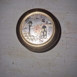 May include: A round gold-toned metal container featuring a circular inlay on the lid. The inlay depicts a person in traditional attire holding a parasol, standing in a landscape with a lantern, cherry blossoms, and a distant pagoda structure.