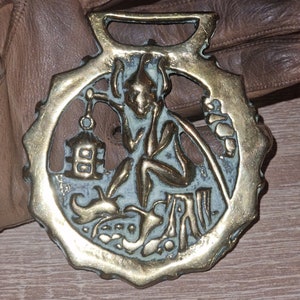May include: A brass horse brass with a detailed design of a figure holding a lantern. The brass is a warm gold color and has a worn patina.