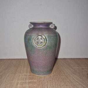 May include: A small, purple and green ceramic vase with a Celtic cross design. The vase has a rounded body and a narrow neck.
