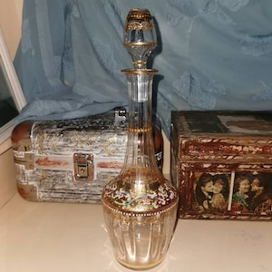 Puede incluir: Una decantador de vidrio transparente con un tapón adornado con oro y diseños florales intrincados en oro, rosa y azul. La decantador tiene un cuerpo texturizado y acanalado.