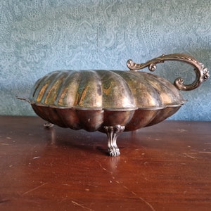 May include: A silver serving dish shaped like a seashell with a handle. The dish has a tarnished finish and sits on four feet.