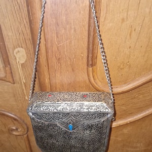 Peut inclure: Sac à main en métal argenté avec des motifs floraux complexes et une chaîne. Le sac rectangulaire est orné de pierres précieuses rouges et bleues. Le sac mesure environ 15 cm de large et 13 cm de haut.