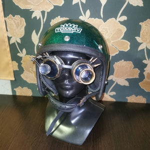 May include: Green motorcycle helmet with a black strap and goggles. The helmet has a white logo that says "TOMMY" with a crown above the word.
