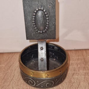 May include: A vintage metal ashtray with a rectangular top and a circular base. The top features an oval beaded design. The base has a gold band with an ornate scroll pattern. The ashtray is made of dark metal.