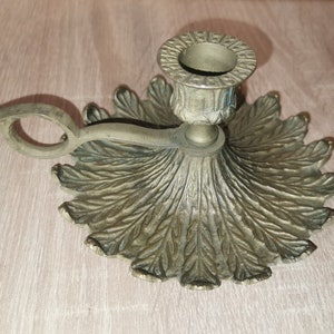 Puede incluir: Un candelero de latón con una base en forma de hoja y un brazo curvo. El candelero tiene un diseño sencillo y un acabado desgastado.