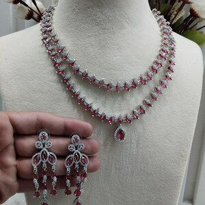May include: A silver necklace with a pink ruby pendant and matching earrings. The necklace is double-layered and features a teardrop-shaped pendant. The earrings are long and dangle. The jewelry is set against a white background.