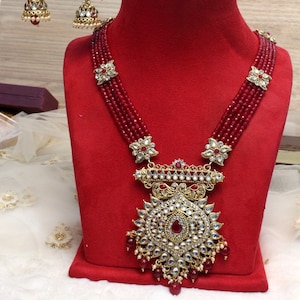 Puede incluir: Un collar de cuentas marrones con un gran colgante adornado con piedras doradas y blancas. El colgante está rodeado de cuentas rojas más pequeñas. El collar se muestra en un soporte de terciopelo rojo. No. 5 - Marrón