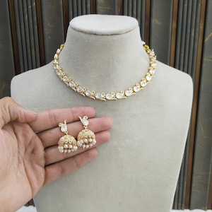 May include: Gold and white necklace and earring set. The necklace is a choker style with a repeating teardrop design. The earrings are bell-shaped with dangling pearls. The set is displayed on a mannequin.