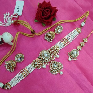 May include: An ornate gold-toned jewelry set featuring a choker necklace, earrings, and a headpiece, all adorned with white pearls and clear gemstones. The set is arranged on a pink surface, with a red rose and a tag visible.